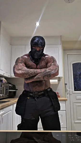 Zade, dark camo balaclava. Arms crossed in kitchen, tactical belt.