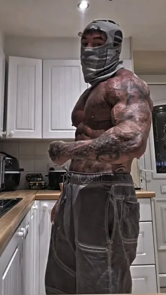 Zade, grey balaclava. Shirtless side-on, arms folded, kitchen.