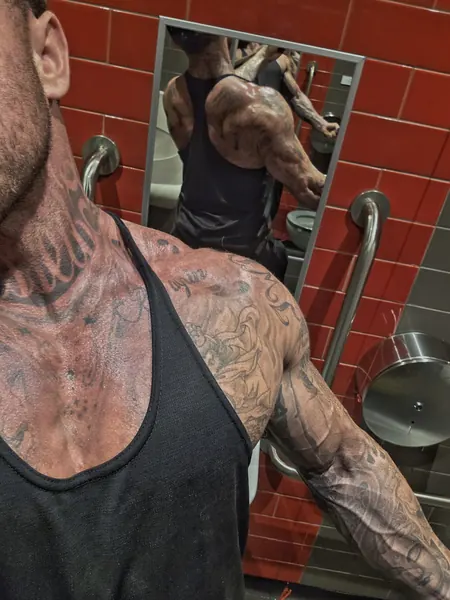 Zade, face hidden. Close-up of tattooed shoulders in black vest, gym mirror.
