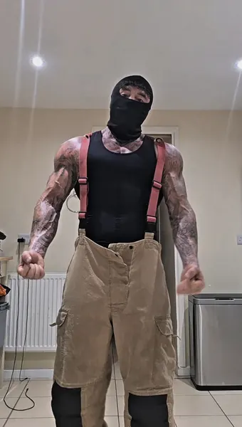 Zade, black balaclava. Black vest and braces, firefighter trousers, low angle.