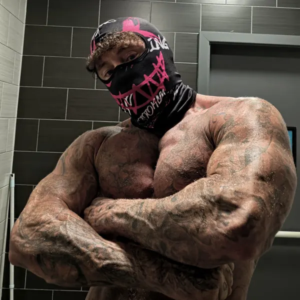 Zade, pink graphic face mask. Arms-crossed close-up in cap and pink graphic gaiter.