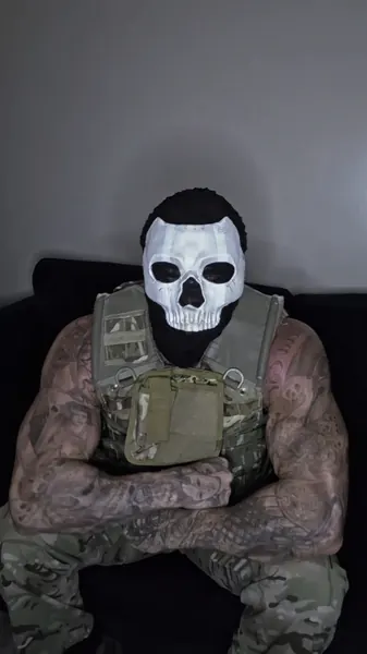 Zade, white skull mask. Seated in plate carrier and camo.