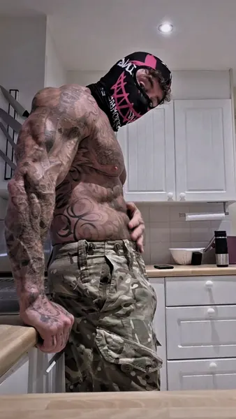 Zade, pink graphic face mask. Leaning on kitchen counter, camo trousers.